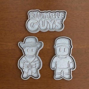 May include: Three grey cookie cutters on a wooden surface. The cutters are in the shape of the Stumble Guys logo and two character figures. One character wears a cowboy hat and the other a cap.