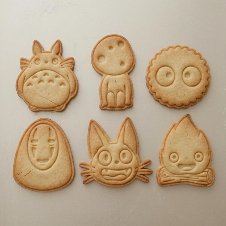 May include: Six baked cookies in various shapes, including a Totoro character, a mushroom-shaped figure, and a round cookie with eyes. The cookies are light brown and appear to be freshly baked, with detailed designs.