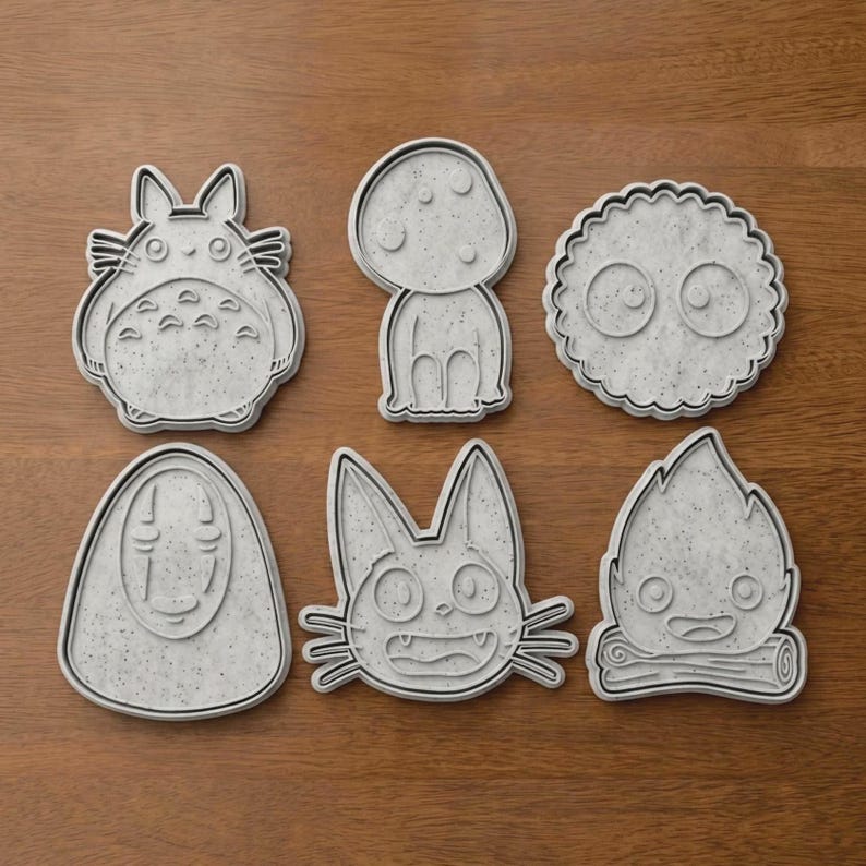 May include: Six gray cookie cutters in various shapes, including a Totoro-like character, a mushroom, a round creature with eyes, a faceless figure, a cat-like creature, and a flame. The cutters are arranged on a wooden surface.