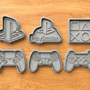 May include: Six grey PlayStation-themed cookie cutters on a wooden surface. The set includes the PlayStation logo, the logo with "PlayStation", a square with controller symbols, and three controller designs.