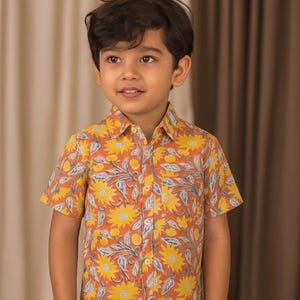 Op de afbeelding: Een shirt met korte mouwen met een bloemenprint. Het shirt heeft een patroon van gele zonnebloemen, blauwe bladeren en oranje fruit op een roestkleurige achtergrond. Het shirt wordt gedragen door een jong kind.