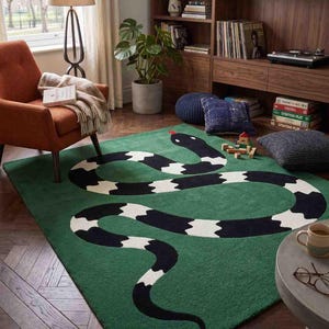 May include: A rectangular area rug featuring a large, stylised snake pattern in black and white against a solid green background. The snake is depicted in a winding, S-shaped position with a small red tongue visible at the head of the serpent.