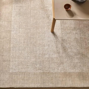 May include: A large rectangular area rug featuring a textured beige and light brown surface with a distinct vertical ribbed pattern. The rug has a solid, smooth border in a matching neutral tone, creating a clean frame around the central design.
