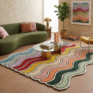 Peut inclure: Un tapis rectangulaire présentant un motif ondulé multicolore composé de rayures bleues, lavande, rouges, jaunes, roses, orange, vert foncé et vert clair avec un bord festonné, posé sur le sol dans une pièce avec un canapé vert et un fauteuil jaune.