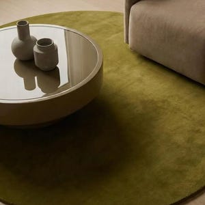 Peut inclure: Un tapis rond vert olive à la texture douce, placé sous une table basse circulaire au plateau marron brillant. Deux petits vases en céramique gris mat sont posés sur la table, accompagnés d'une partie d'un fauteuil rembourré beige.