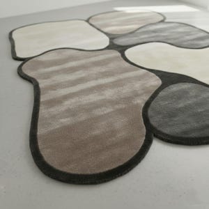 May include: A modern area rug with an organic, pebble-like design. The rug features various shades of gray, beige, and brown, with a dark gray border. The soft, textured surface adds visual interest and warmth to any room. This unique rug is a stylish home decor accent.