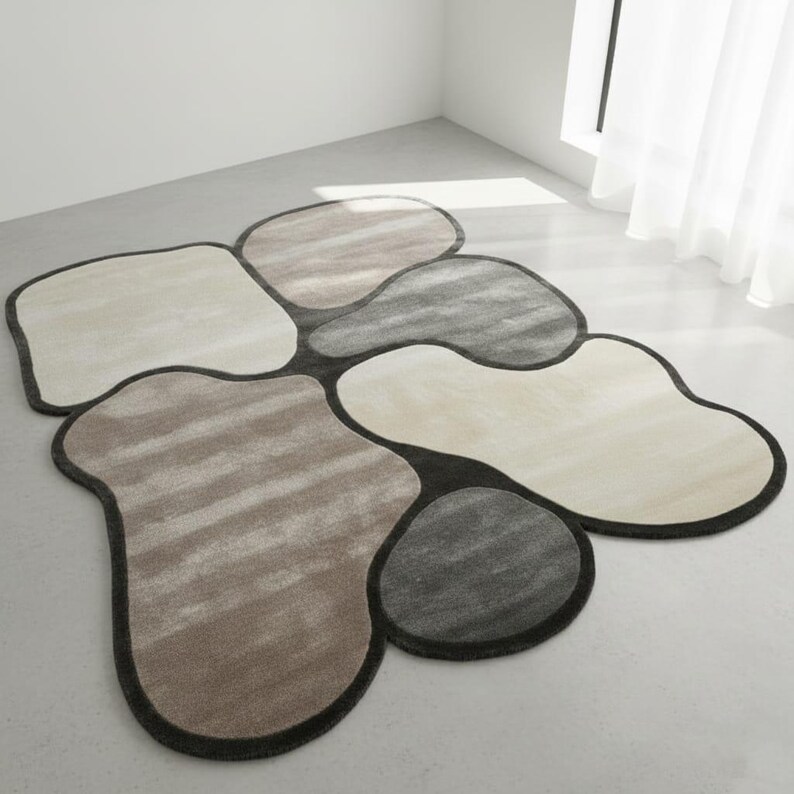 May include: A modern area rug with an abstract design. The rug features several irregularly shaped sections in shades of beige, gray, and brown, outlined with a dark gray border. The rug is made of a soft, textured material.