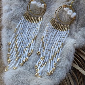 May include: A pair of handcrafted earrings featuring gold-toned hoops with white and clear stone accents. Long strands of white and gold beads cascade from the hoops, creating a fringe effect. The earrings are displayed on a textured, light-colored surface.