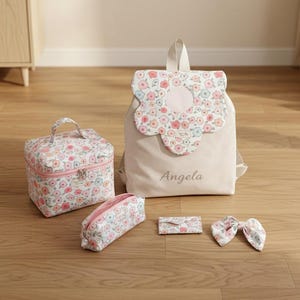 Puede incluir: Un conjunto de accesorios a juego, que incluye una mochila, un neceser, un estuche, un tarjetero y un lazo. Los artículos presentan un estampado floral en rosa, blanco y azul sobre un fondo crema. La mochila tiene una solapa con forma de flor y el nombre "Angela" impreso.