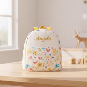 May include: A small, white and floral-patterned backpack with yellow straps. The backpack has the name "Angela" embroidered in gold. The lower portion of the backpack features a colorful floral design with hearts and suns.