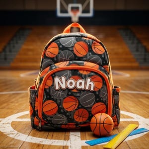 May include: A basketball-themed backpack with the name "Noah" printed on the front. The backpack features a pattern of orange and gray basketballs on a black background, with orange trim. A small basketball and two rulers are next to the backpack.