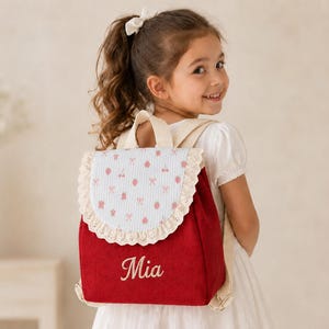 May include: A red corduroy backpack featuring a light blue and white striped flap with a pink cherry and bow pattern. The flap is trimmed with cream-colored lace. The name Mia is embroidered in white script on the front of the red backpack body.
