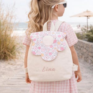 Könnte beinhalten: Ein cremefarbener Rucksack mit einem Blumendesign auf der Klappe, getragen von einem Kind. Der Rucksack hat den Namen "Lina" auf der Vorderseite gestickt. Das Kind trägt ein rosa Vichy-Kleid und eine Sonnenbrille.