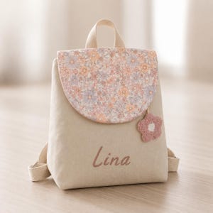 May include: A beige backpack with a floral patterned flap and a pink crocheted flower charm. The name "Lina" is embroidered in pink on the front. The backpack has a top handle and adjustable straps.