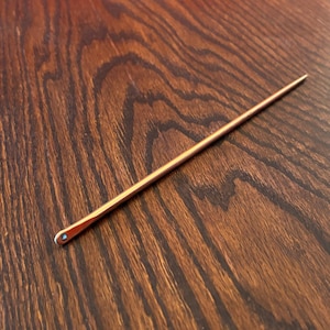 May include: A slender, copper-colored hair pin with a pointed end and a small, decorative accent at the opposite end. The pin is set against a dark wood grain surface.