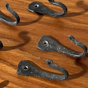 May include: Several small, black metal hooks with a rustic, hammered appearance are arranged on a wooden surface. Each hook has a curved design and a small hole for mounting. The hooks are likely for hanging items.