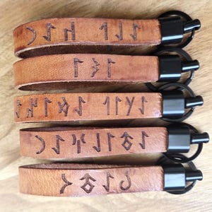 Könnte beinhalten: Fünf braune Leder-Schlüsselanhänger mit schwarzen Metallbeschlägen. Jeder Schlüsselanhänger hat eingravierte Runen. Das Leder hat ein natürliches, abgenutztes Aussehen. Die Schlüsselanhänger sind in einer ordentlichen, parallelen Reihe angeordnet.