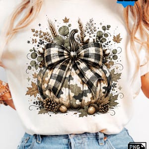 May include: A cream-coloured t-shirt featuring a graphic of a pumpkin with a camouflage pattern and a black and white buffalo plaid bow. The pumpkin is surrounded by autumn foliage, wheat stalks, cotton bolls, pinecones, and acorns.