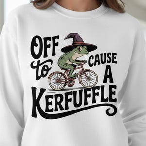 May include: White sweatshirt with a whimsical graphic of a frog riding a bicycle and wearing a witch's hat. The text reads "Off to Cause a Kerfuffle" in black script.