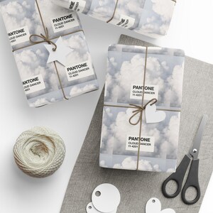 May include: Gift-wrapped presents with cloud-patterned paper and twine, adorned with white star and heart-shaped tags. The paper features the text "PANTONE CLOUD DANCER 11-4201". A spool of twine, scissors, and blank tags are also visible.