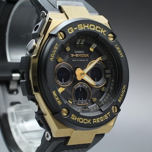 May include: A G-Shock watch with a black and gold-tone design. The watch features a black dial with gold hands and markers, and the text "G-SHOCK" and "SHOCK RESIST" are visible. The watch has a black strap.