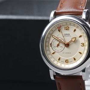 May include: An Oris Automatic wristwatch with a silver-tone stainless steel case and a brown leather strap. The cream-colored dial features gold-tone hands, Arabic numerals at 3, 6, and 12, a sub-dial, a date pointer hand, and the text Oris Automatic.