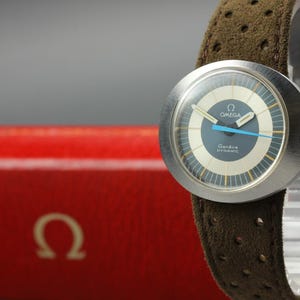 May include: An Omega Genève Dynamic watch with a round silver-tone case, a blue and cream dial featuring the text OMEGA Genève DYNAMIC, and a bright blue second hand. The watch is fitted with a brown perforated suede leather strap.