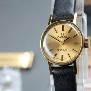 Pode incluir: Um relógio de pulso Omega Genève em tom dourado com mostrador redondo dourado, marcadores de hora pretos finos, ponteiros pretos e pulseira de couro preta. O mostrador exibe o logotipo Omega, o texto OMEGA e Genève e Swiss Made abaixo.