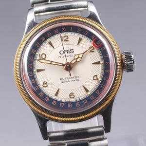 Może przedstawiać: Zegarek automatyczny Oris w stylu vintage z bransoletą ze stali nierdzewnej w kolorze srebrnym. Kremowa tarcza zegarka ma złote wskazówki i znaczniki. Posiada niebieską i złotą ramkę z wskaźnikiem daty oraz napis "ORIS 17 JEWELS AUTOMATIC SWISS MADE".