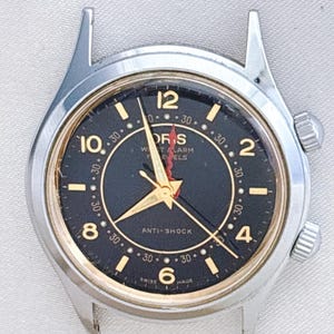 Może przedstawiać: Zegarek naręczny w stylu vintage z czarną tarczą i złotymi wskazówkami i cyframi. Zegarek ma srebrną kopertę i dwie koronki do nakręcania. Tarcza zegarka zawiera tekst "ORIS", "W. ALARM" i "JEWELS". Zegarek ma również tekst "ANTI-SHOCK" i "SWISS MADE".