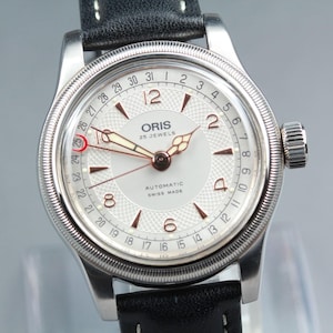 May include: Silver wristwatch with a white face, displaying the brand name "ORIS" and "25 JEWELS". The watch has a black leather strap and a date display. The watch hands are gold-toned.
