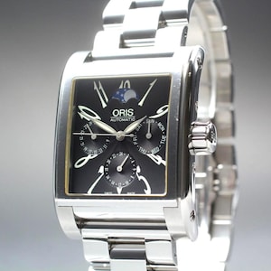 May include: A silver-toned Oris automatic watch with a rectangular black face. The watch features a moon phase indicator, day and date subdials, and a stainless steel band. The watch face displays the brand name and "automatic".
