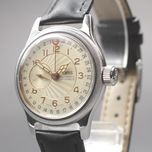 May include: A silver wristwatch with a cream face and black leather strap. The watch face displays Arabic numerals, a subdial, and the brand name "ORIS". The watch has a textured bezel and a crown. The watch is approximately 3.5 cm in diameter.