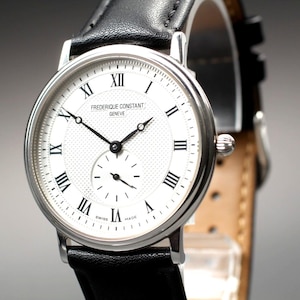 Puede incluir: Un reloj de pulsera plateado con esfera blanca y números romanos negros. El reloj presenta un pequeño dial de segundos y agujas negras. La correa de cuero negro complementa el diseño clásico del reloj. El texto "FREDERIQUE CONSTANT GENEVE" es visible en la esfera.