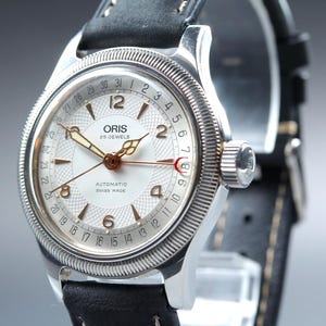 Puede incluir: Un reloj de pulsera plateado con esfera blanca y correa de cuero negro. La esfera muestra la marca "ORIS", el texto "25 JEWELS" y "AUTOMATIC SWISS MADE". Tiene un indicador de fecha y un bisel texturizado. Aproximadamente 40 mm de diámetro.