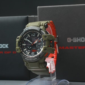 Op de afbeelding: Een olijfgroen G-Shock horloge met een zwarte wijzerplaat en rode accenten. Het horloge wordt gepresenteerd op een transparante standaard. Het horloge staat naast een zwarte doos met de woorden "G-Shock" en "Master of G" in wit en rood. Het heeft een digitale display.