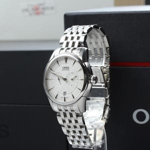 May include: An Oris automatic wristwatch with a round silver-toned dial, silver hands, and hour markers. The watch features a multi-link stainless steel bracelet and a date window at the six o'clock position. The Oris logo is printed on the dial.
