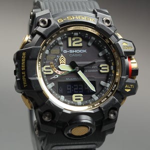May include: Black and gold G-Shock watch with a digital display showing the time as 9:23. The watch has a black band and a round face with gold accents. The watch face includes the text "G-SHOCK CASIO".