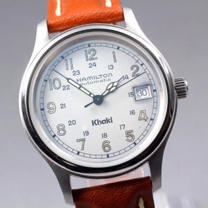 Può includere: Un orologio automatico Hamilton Khaki argentato con quadrante bianco, numeri neri e datario. L'orologio ha un cinturino in pelle marrone con cuciture bianche.