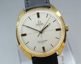 Può includere: Un orologio da polso automatico Omega Seamaster Cosmic con cassa color oro, quadrante bianco argento e indici neri. L'orologio presenta un cinturino in pelle nera, una corona sul lato destro e la scritta OMEGA AUTOMATIC Seamaster COSMIC.