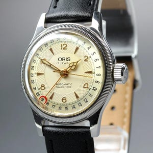 May include: An Oris automatic wristwatch with a silver-tone case, a textured cream dial, and a black leather strap. The dial features gold-tone hands, Arabic numerals at 12, 2, 4, 6, and 8, and a date pointer hand indicating the 19th day.
