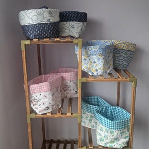 May include: A wooden shelving unit holds several fabric storage baskets. The baskets have patterns such as anchors, bicycles, and gingham. The colour scheme includes navy, white, pink, blue, and yellow. The baskets are of different sizes and shapes.