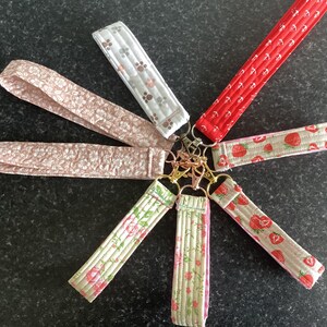 May include: A selection of fabric wrist keychains in diverse patterns, including floral, paw prints, and strawberries. The keychains are attached to gold-coloured metal clasps. The fabrics feature shades of pink, red, white, and green.