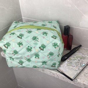 May include: A quilted cosmetic bag with a light green background and a pattern of green frogs. The bag has a green zip. Other beauty products are on the countertop. The bag measures approximately 20 cm x 20 cm.