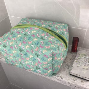 May include: A mint green quilted cosmetic bag patterned with butterflies in pink, blue, and yellow. It has a green zip. Beside it are a red lipstick and a decorative silver rose in a box.