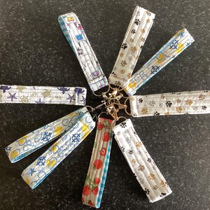 May include: A selection of fabric wrist keychains with diverse patterns, including nautical themes, paw prints, and fruit designs. Each keychain is attached to a gold-coloured metal clasp. The keychains are made of quilted fabric and feature a variety of colours.