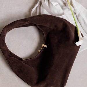 Può includere: Una borsa a spalla in pelle scamosciata marrone scuro con chiusura a zip dorata. Un sacchetto bianco con coulisse e una calla bianca con stelo verde sono sullo sfondo. La borsa ha una forma curva a mezzaluna.