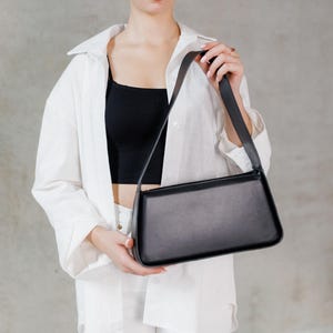 Könnte beinhalten: Eine schwarze, rechteckige Umhängetasche mit langem Riemen. Die Tasche ist aus glattem Material gefertigt und hat ein minimalistisches Design. Die Tasche wird von einer Person gehalten, die ein weißes Hemd und ein schwarzes Oberteil trägt.