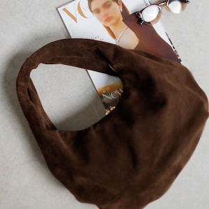 May include: A brown suede hobo bag with a single shoulder strap. The bag is resting on a magazine with a blurred image of a woman on the cover, and a pair of sunglasses.