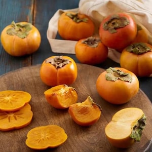 May include: A collection of bright orange persimmons, some whole with green stems and others sliced into rounds and wedges, arranged on a round wooden cutting board alongside a cream-colored fabric bag on a dark blue wooden surface.